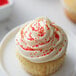 A cupcake with white frosting and red nonpareils on a plate.