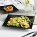 A black square plate with salad and fruit on a table.