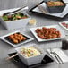 A table with Acopa black square melamine plates of food, including a salad, rice, and a clear container with yellow liquid.