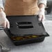 A woman in gloves using a Vigor black polycarbonate food pan lid to cover a black container of food.