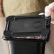 A person holding a black Vigor polycarbonate food pan lid over a container of food.