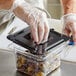 A person wearing a plastic glove in a close-up opening a Vigor black plastic food container with olives.