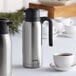 A silver stainless steel Thermos carafe with a black twist top.
