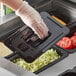A hand in a plastic glove putting food into a Vigor 1/4 size black polycarbonate food pan with a lid.