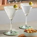 Two Schott Zwiesel martini glasses with olives on the table with a bowl of nuts.