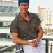 A man wearing an olive green Uncommon Chef cook shirt with a white apron and chef's hat smiling at the camera.