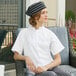A woman sitting in a professional kitchen wearing a white Uncommon Chef cook shirt with a black striped hat.