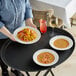 A woman holding a black oval non-skid serving tray with a plate of pasta and a glass of white wine.