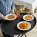 A woman serves pasta with shrimp and soup on a black non-skid oval serving tray.