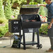 A man using a Backyard Pro wood-fired pellet grill to barbecue meat on a table in an outdoor setup.