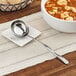 A solid stainless steel ladle in a bowl of soup on a table with silverware.