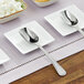 A stainless steel scoop spoon with a mirror finish in a bowl of sugar cubes on a table with a plate of spoons.