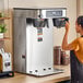A woman standing in front of a Bunn stainless steel coffee maker on a counter.