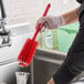 A person in rubber gloves washing dishes in a professional kitchen sink with a red Carlisle Sparta bottle cleaning brush.