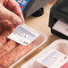 A hand applying a white pre-printed safe handling label to a package of sausages, with a label printer and another labeled meat package nearby.