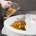 A hand using a clear scoop to pour multicolored pasta from a white container.