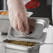 A person wearing plastic gloves putting food into a Carlisle stainless steel slotted steam table pan with a spoon.