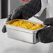 A gloved hand holding a Carlisle stainless steel steam table pan filled with macaroni and cheese.