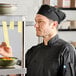 A black chef skull cap or pill box hat with a mesh top, worn by a chef in a kitchen setting.