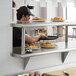 A man in a black hat standing behind a Regency stainless steel pass-through shelf with an overshelf displaying plates of food.