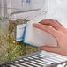 A hand holding a white Puracycle melamine foam sponge over a white container of beans.