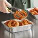 A person using tongs to pick up a fried onion ring from a tin on a counter.