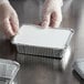 A person in gloves holding a Choice deep oblong foil container with a white board lid.