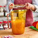 A woman using a Cambro measuring cup to pour orange juice from a glass jar.