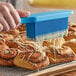 A person using a blue Ateco silicone bristle brush to baste a cinnamon roll.