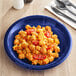 A navy blue plastic plate with pasta and tomatoes on it, with a black plastic spoon and fork on a napkin.