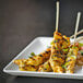 A Front of the House square porcelain plate with skewers of chicken and rice on a table.