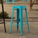 A turquoise metal Lancaster Table & Seating outdoor barstool on a stone surface.