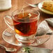 A glass cup of tea with a Steep Cafe By Bigelow English Breakfast Decaffeinated Tea bag on a saucer.