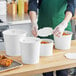 A person wearing a plastic glove putting food in a white Choice food bucket with a lid.