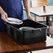 A man in a blue uniform placing plates and utensils in a black Vollrath bus tub.