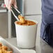 A person using metal tongs to serve fried chicken from a white Choice food bucket.