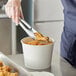 A person using tongs to put food in a Choice white food bucket.