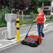 A woman in a red shirt pushing a Bissell Commercial outdoor power sweeper on a sidewalk.