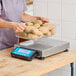 A digital receiving scale with a stainless steel platform and a blue control panel, being used to weigh a container of potatoes.