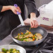 A person using a Choice aluminum frying pan with a purple handle to cook food.