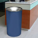 An indigo Ex-Cell Kaiser Premier Series steel round waste receptacle with a textured surface sits in front of a glass door.