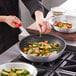 A person cooking food in a Choice non-stick fry pan with a red silicone handle.