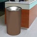 A brown Ex-Cell Kaiser steel round waste receptacle with a textured surface in front of a reception desk.