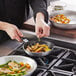 A person cooking food in a Vollrath Arkadia aluminum non-stick fry pan with a black silicone handle.