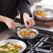 A person cooking food in a Vollrath aluminum fry pan with a black silicone handle on a stove.