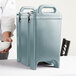 A person holding a bowl of food next to a blue Cambro insulated soup carrier.