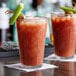 Two Duralex highball glasses filled with bloody mary on a table with celery and olives.