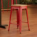 A Lancaster Table & Seating red metal backless bar stool with a natural wood seat.