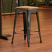 A Lancaster Table & Seating black metal stool with a natural wood seat on a wood floor.