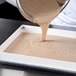 A person pouring brown liquid from a bowl into a white fiberglass tray using a fiberglass sheet pan extender.